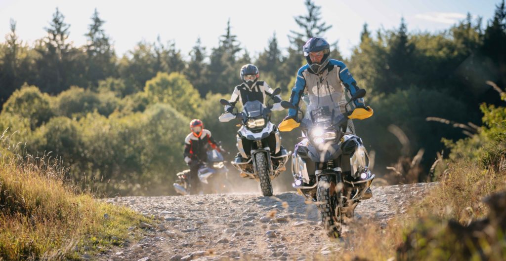 Drei Motorradfahrer auf BMW GS Adventure Motorrädern fahren auf einem steinigen Waldweg. Der vorderste Fahrer trägt einen grauen und blauen Anzug und einen blauen Helm. Der zweite Fahrer trägt einen weißen Anzug und einen dunklen Helm. Der dritte Fahrer ist etwas weiter entfernt und trägt einen orangefarbenen Anzug und einen roten Helm. Die Motorräder sind mit Gepäck ausgestattet. Der Weg führt durch einen Wald mit Bäumen und Büschen unter einem hellen Himmel.