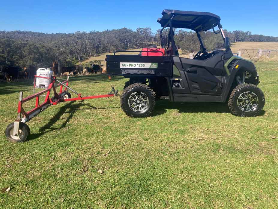 Ein grüner Geländewagen zieht auf einer Weide eine rote Sprühvorrichtung vor einer grasenden Rinderherde im Hintergrund.